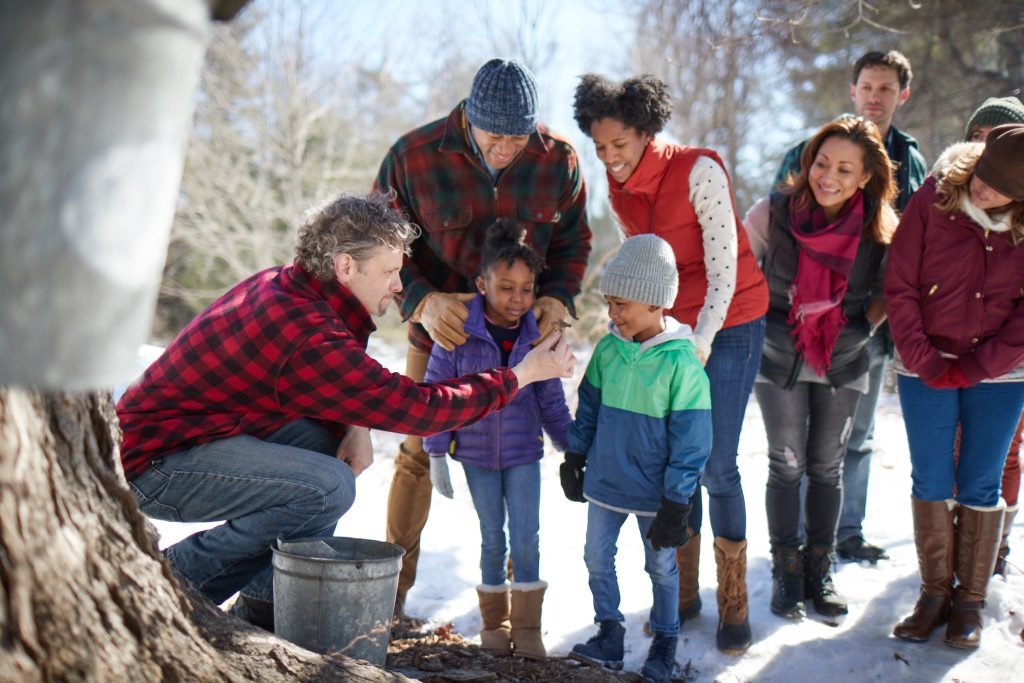 A family leaning in to a man in a red plaid shirt demonstrating how sap is collected in buckets from maple trees.