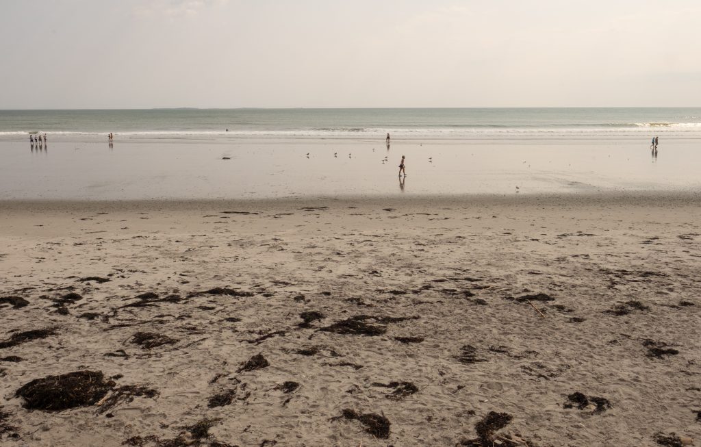 A long, wide gray beach with people walking in the surf in the distance.