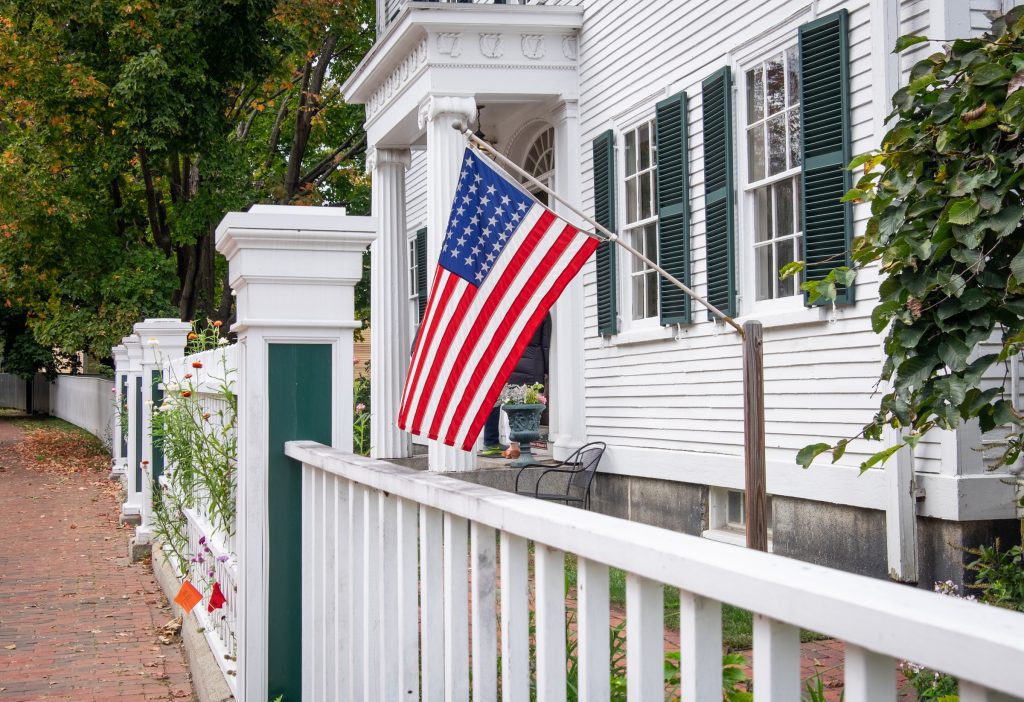 A white fence in front of a white historic building with green shutters, an American flag hanging out front.