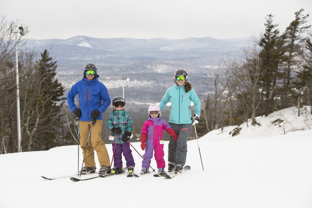 A family with a dad, mom, and two little kids standing on top of a ski mountain in brightly colored snowsuits.