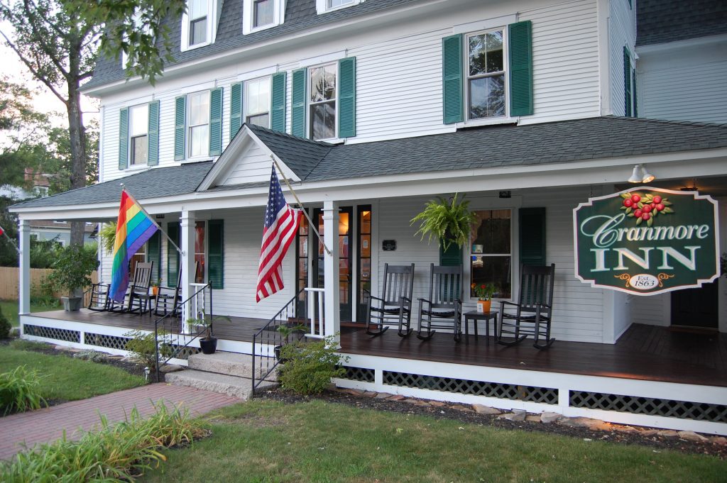 A white B&B with green shutters and a wraparound porch with a rainbow flag, an American flag, and a sign that reads Cranmore Inn.