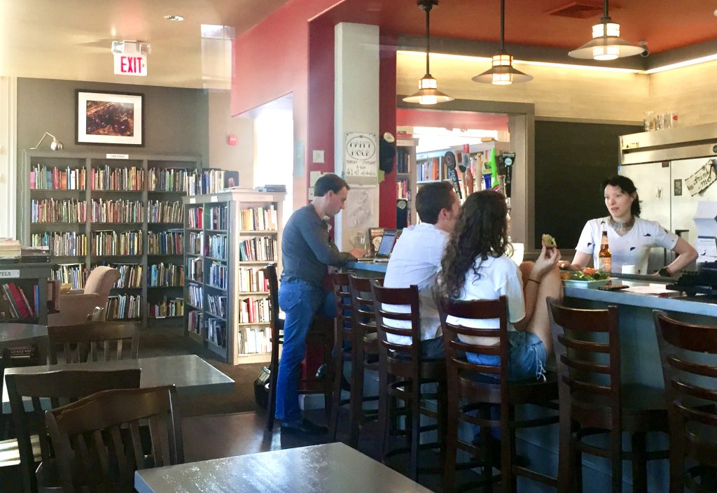 A bar clearly set in a bookstore, people sipping beers and chatting with the bartender.