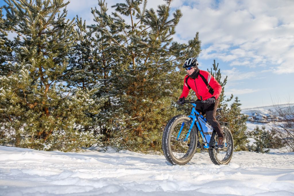 A man in a red jacket riding a fat tire bicycle through a snowy forest.