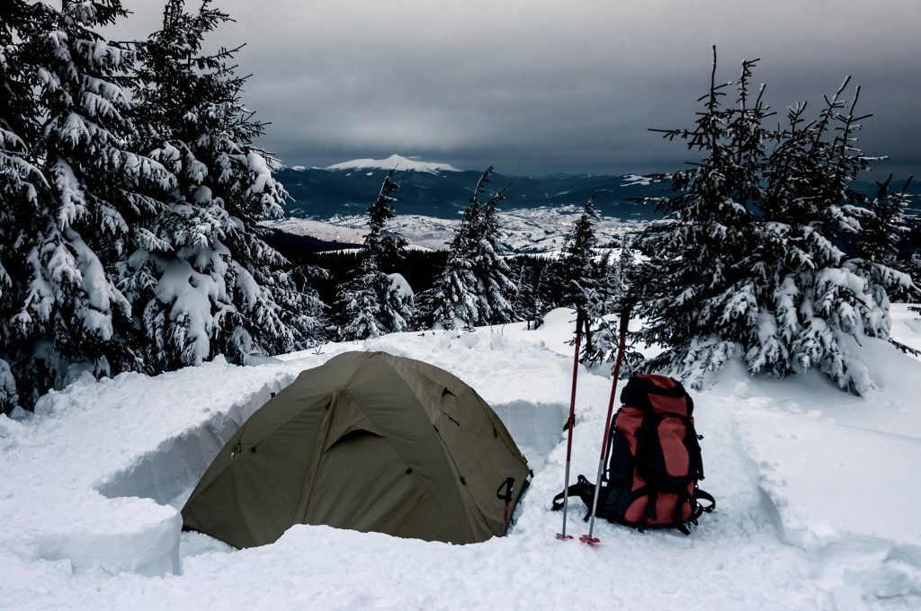 A tent and backpack on a snowy mountain surrounded by evergreen trees.