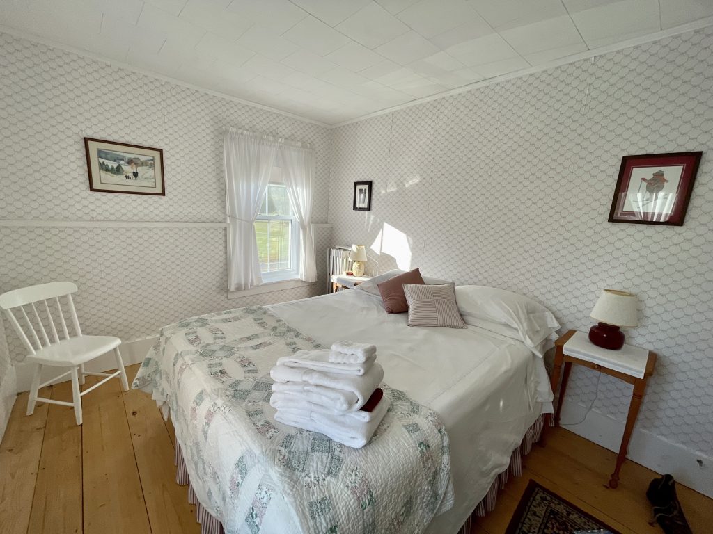 A simple bedroom in a guesthouse, all white and gray, with heavy linens and a pile of white towels on the bed.