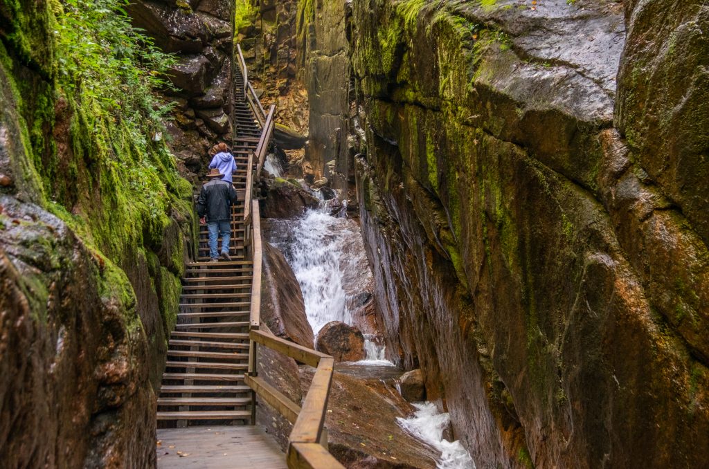 A man and woman climbing up a wooden staircase next to a rushing waterfall between two rock walls.