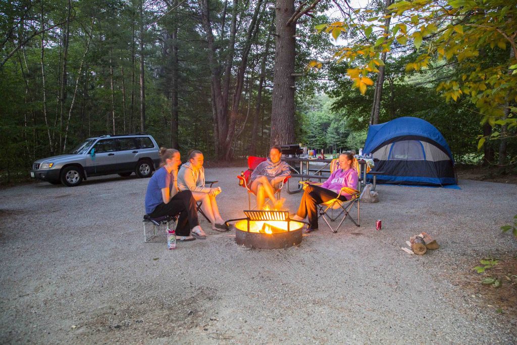 Four young women sitting in camping chairs around a fire, a tent in the background.