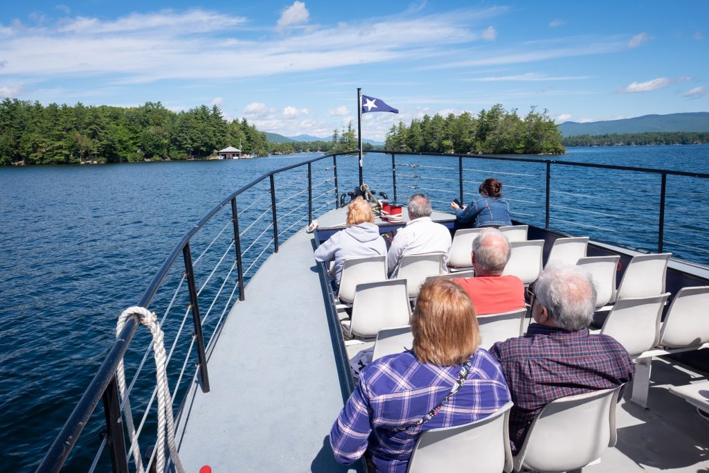 Several seniors sitting in seats in the front of a boat on Lake Winnipesaukee, sailing toward forested islands, blue mountains in the background.
