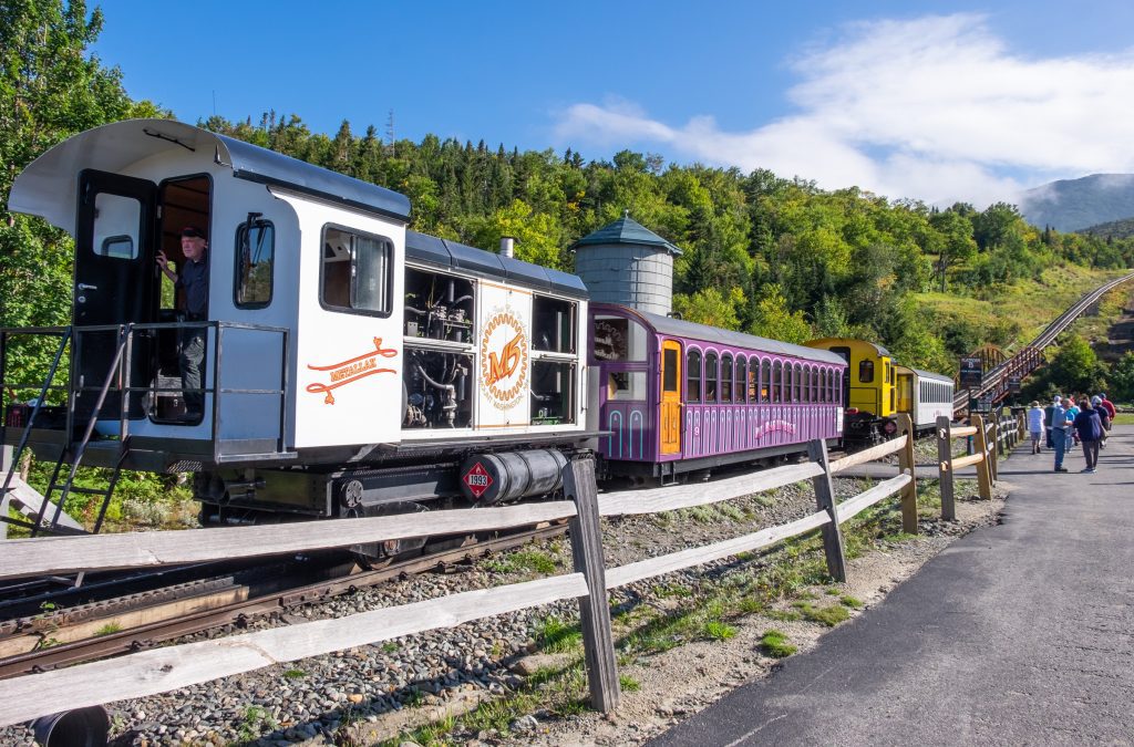 The old-fashioned white, purple and yellow Mount Washington Cog Railway about to leave and climb up a steep mountain.