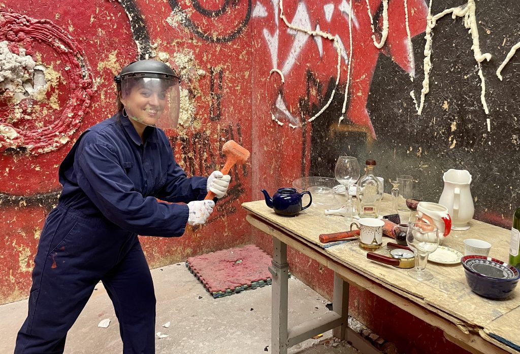 Editor Kate standing in a blue jumpsuit and helmet with face shield, holding a hammer and ready to smash a table full of glass things at the Rage Cage.