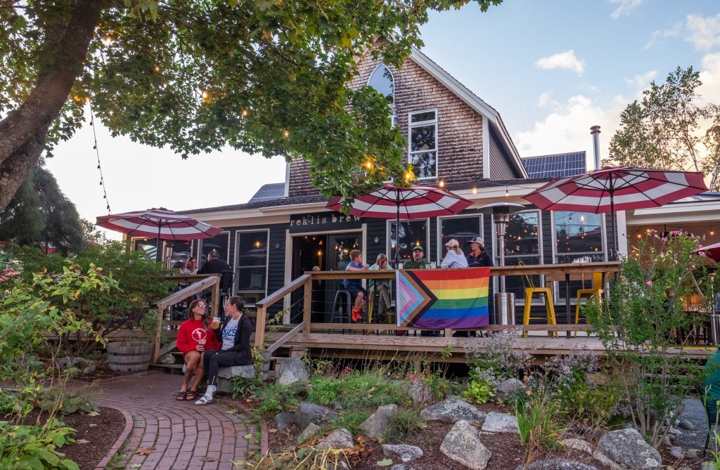 A big shingled house with a front porch, garden, Pride flag hanging in the front, and two girls sitting on the steps drinking beers.