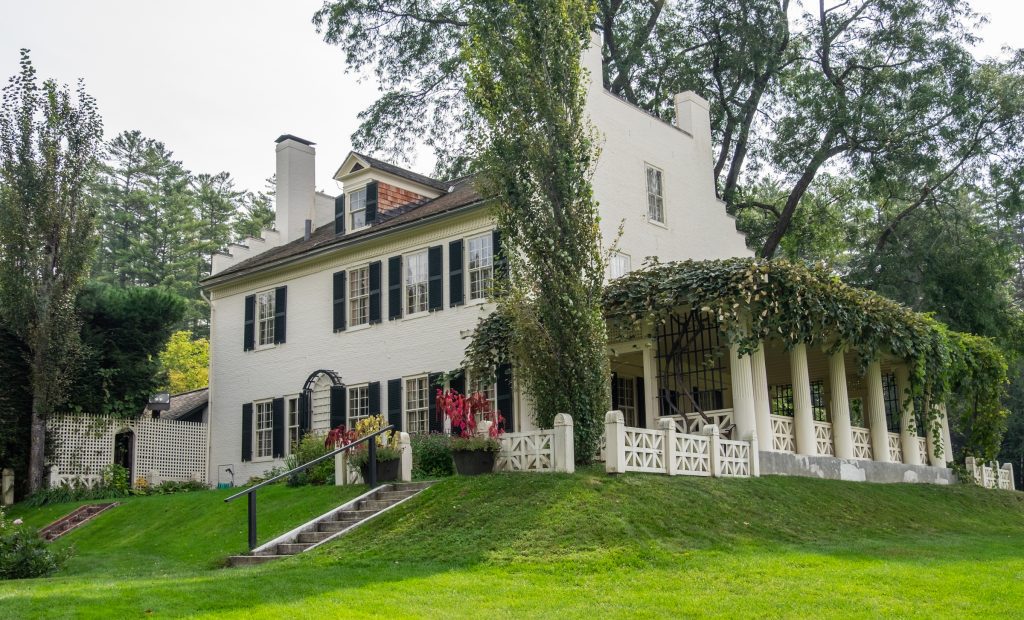 A beautiful white house with black shutters. To one side is a porch with white columns, all covered in green ivy.