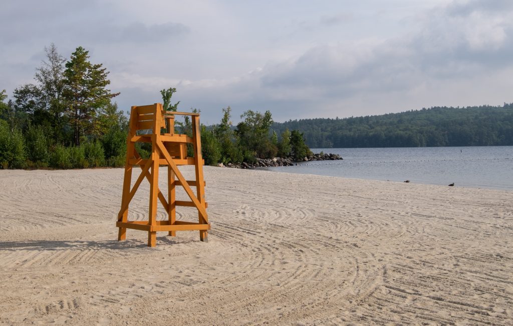 A solitary tall orange lifeguard's chair in the middle of a recently raked sandy beach on the edge of a gray blue lake underneath a cloudy sky.