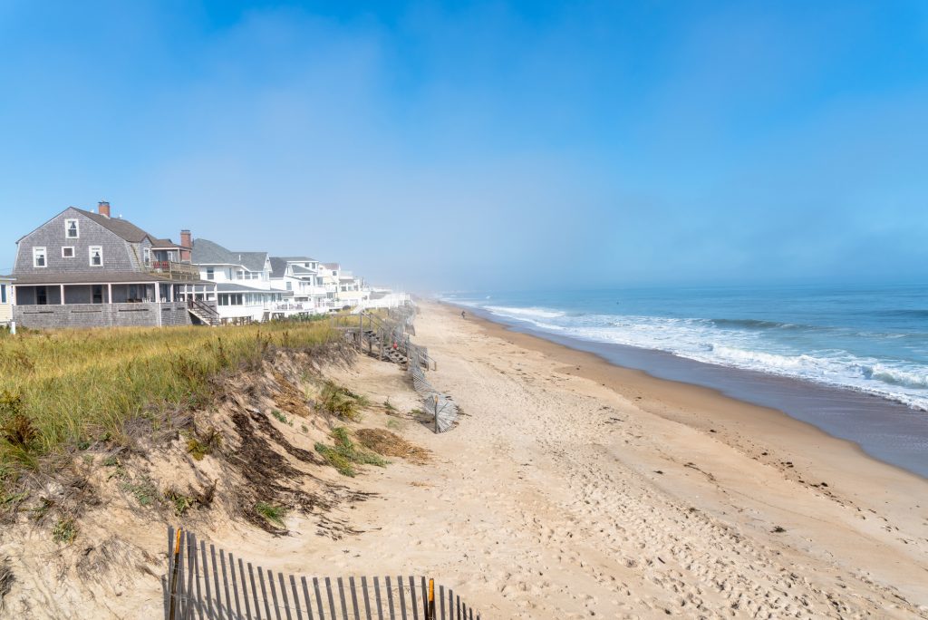 Tall sand dunes topped with grass leading to giant shingled beach mansions. The water is bright blue with big waves.