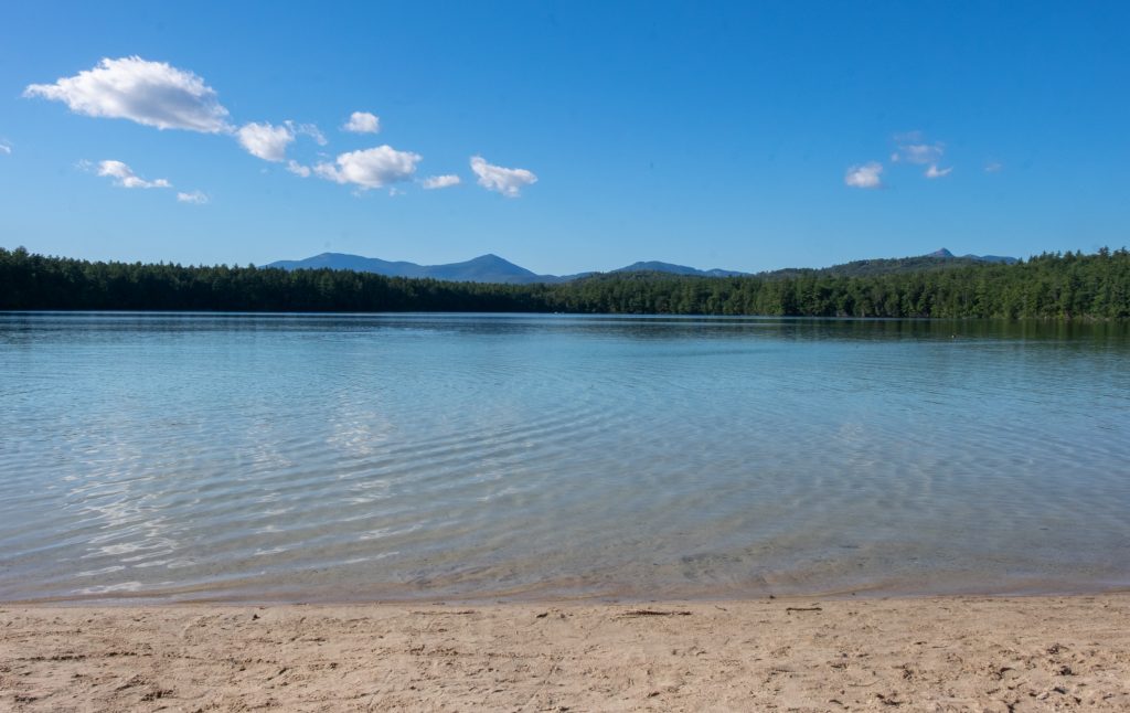 A small piece of sand leading to a lake so calm you can't see a single wave, with bright blue water and trees and bright blue mountains in the background.
