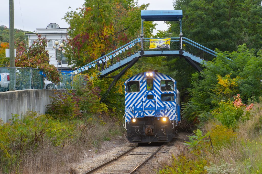 A blue train chugging along a track underneath a pointed footbridge.