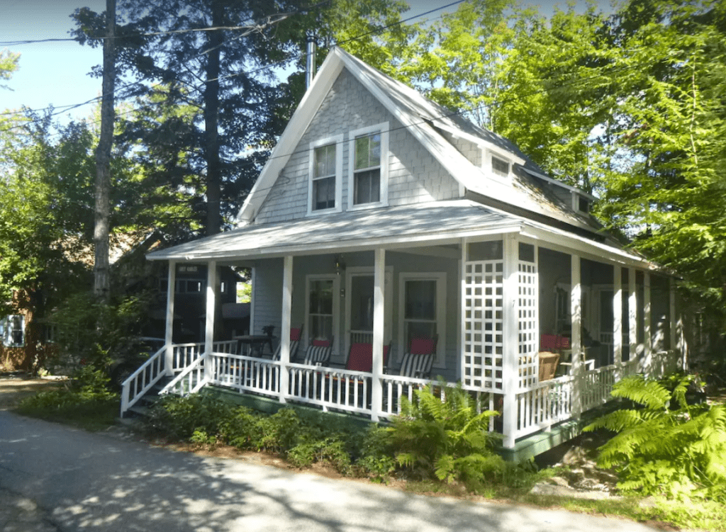 A street view of a gray cottage with a wrap around porch in front of a forest