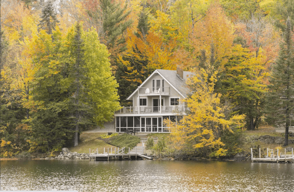 A gray lakefront cottage surrounded by fall foliage, with a private dock