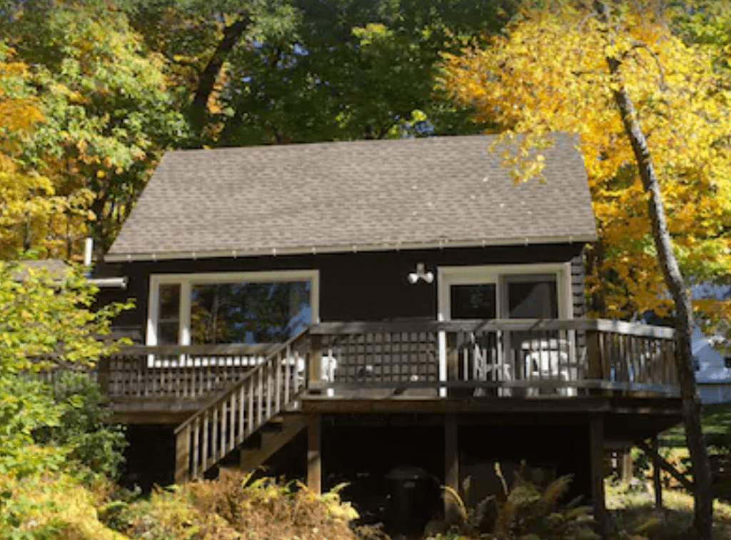 A brown cottage with a wraparound porch in front of fall foliage