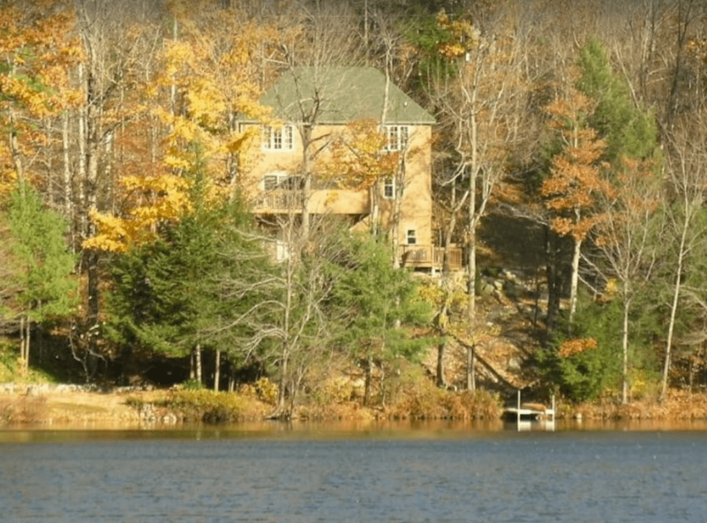 A view of a two story cottage across a lake, partially hidden by some fall foliage