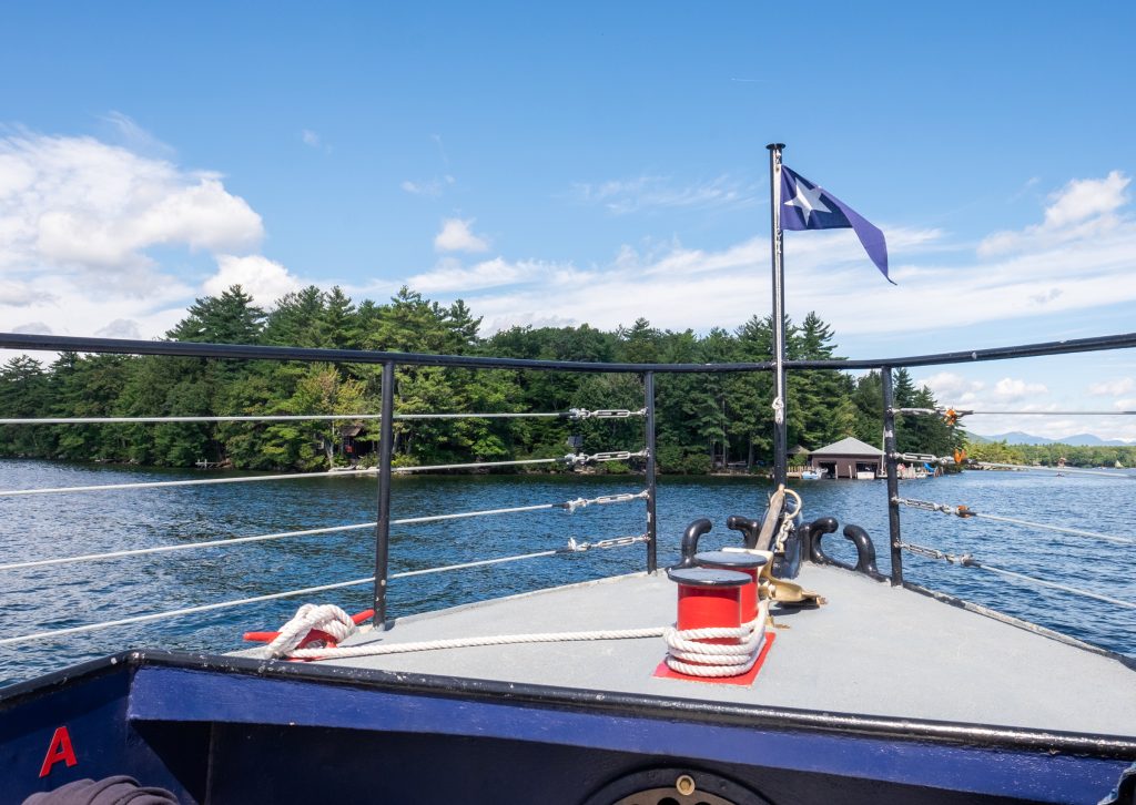 The front of a boat flying a navy blue flag with a single star, cruising toward forested islands on the lake.