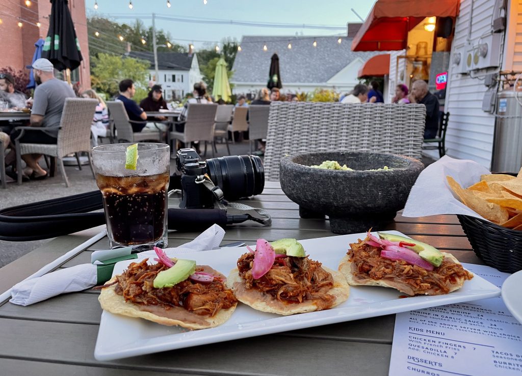 A table at an outdoor restaurant. On a plate are three tacos topped with roasted pork and pink pickled onions.