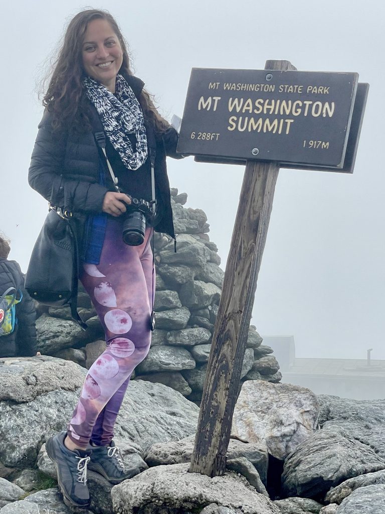Editor Kate smiling, standing next to the sign that reads Mt. Washington Summit, 6288 feet, 1917 meters. She has long curly dark hair and wears a black puffy jacket, black and white scarf, and pink leggings with moons on them, and gray sneakers. She carries a DSLR camera.
