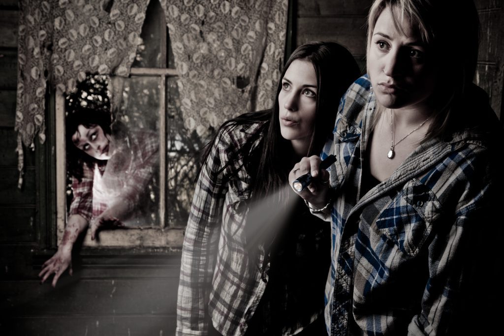 Two women in plaid looking around the corner, with a spooky creature breaking through the window in the background