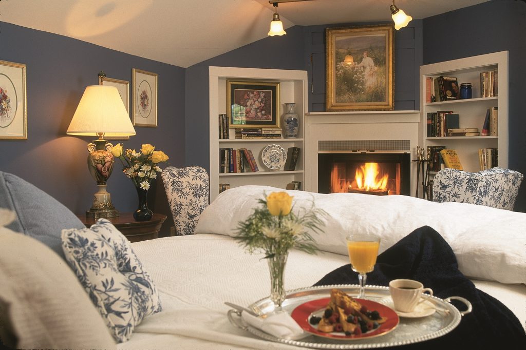 A cozy blue hotel room, with breakfast on the bed and a fireplace blazing in the corner