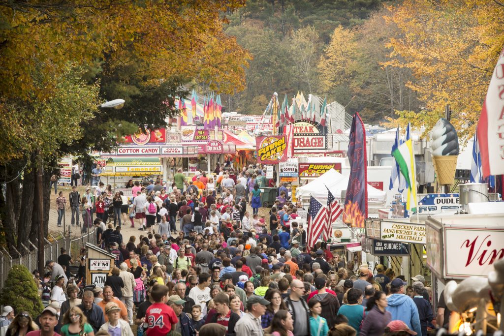 Aerial shot of a large crowd at a New Hampshire Fall festival, with fair food stands and fall foliage in the background