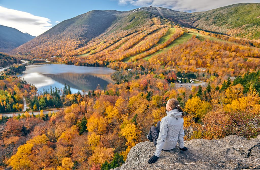 A woman sitting on the edge of a rock ledge, facing a still blue lake surrounded by mountains covered with orange trees.