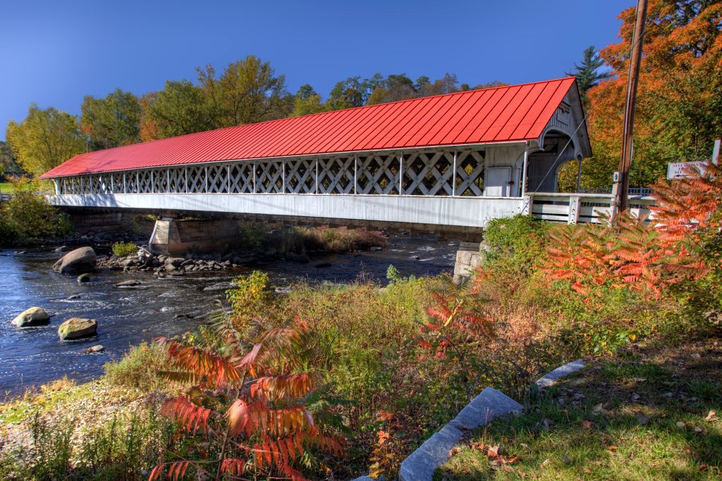 A white covered bridge with a red roof crossing a river, surrounded by red and yellow trees on a fall day.