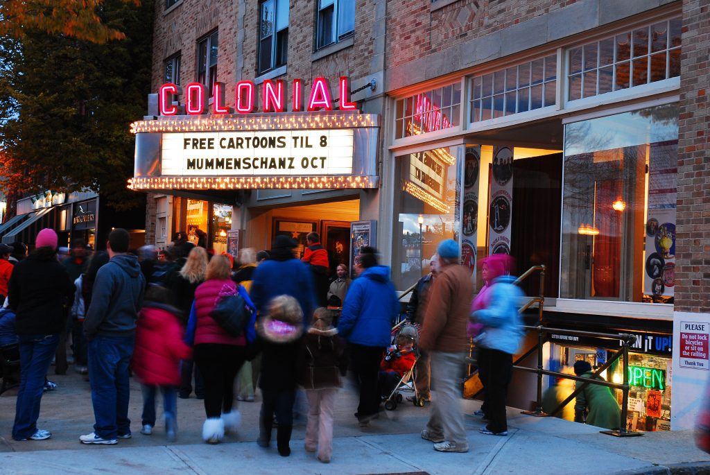 People waiting outside a theater with a pink neon sign reading Colonial and a marquee reading Free Cartoons Til 8.
