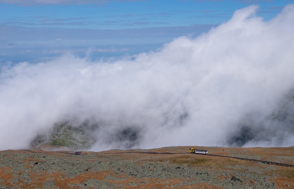 A tiny-looking train heading down the edge of a mountain into a cloud.