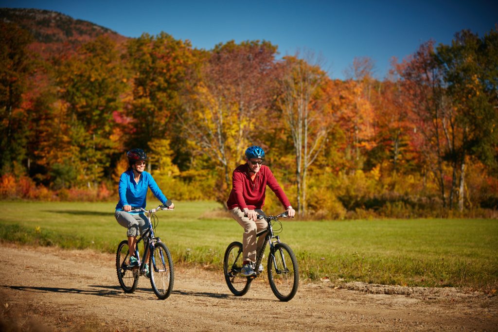 A man and a woman riding bikes along a path, bright yellow and orange trees behind them.