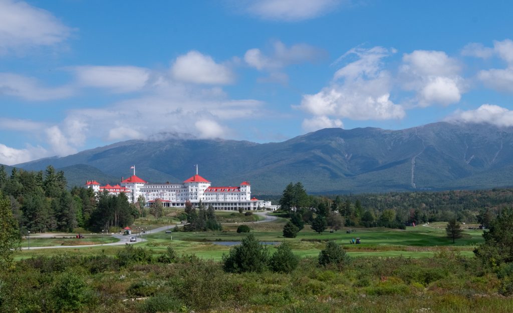 A majestic white hotel with a red roof set amongst the mountains. You can faintly see a rail track in the distance on one of the mountains.