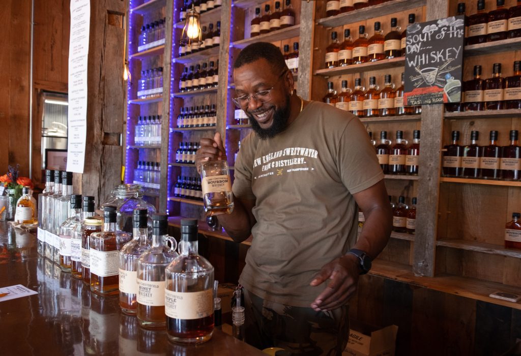 Kenny, a bearded man in glasses and a New England Sweetwater t-shirt, holding up a bottle of bourbon amid his other bottles and smiling widely. Behind him a sign reads "Soup of the Day: Whiskey."