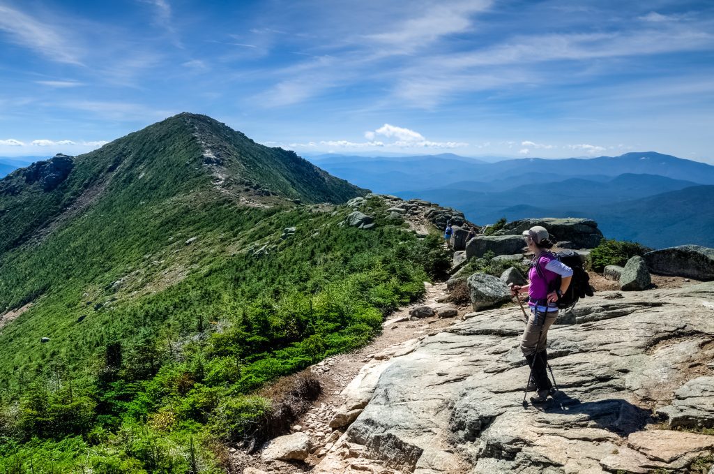 A woman with hiking poles pauses to look at a trail heading up a narrow ridge.