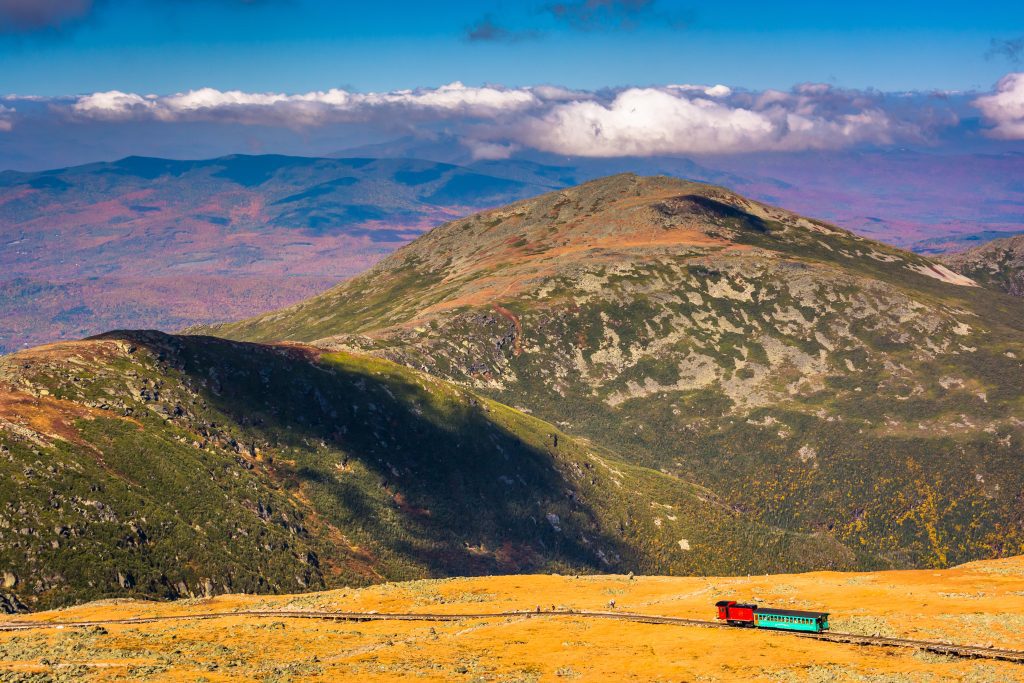 A tiny train heading down the mountainside, surrounded by more mountains covered in orange and gold foliage.