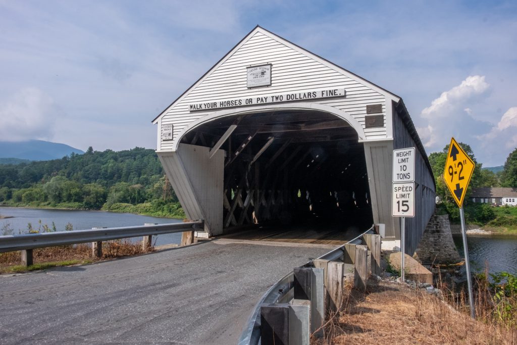The front of a covered bridge. Above the opening a sign reads "Walk your horses or pay two dollars fine."
