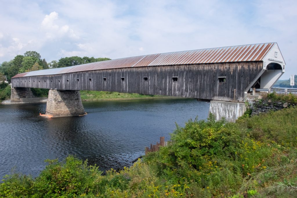 A long wooden covered bridge spanning a wide river in a rural area.
