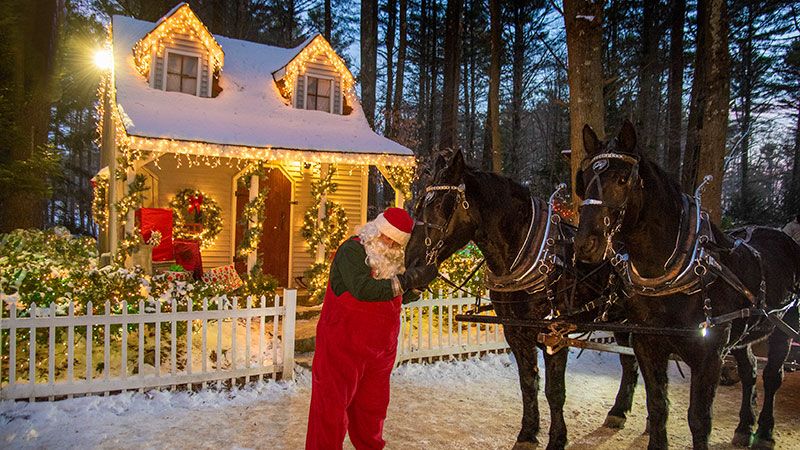 Santa tending to horses outside a cottage lit up with Christmas lights.