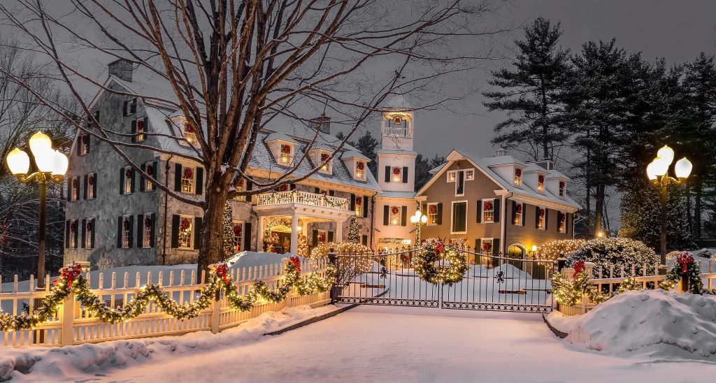 A large white old-fashioned inn covered in Christmas lights and garlands under fresh snowfall at night.
