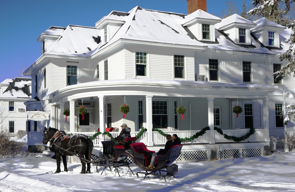 A big white inn covered in Christmas garlands with a horse-drawn sleigh in the snow in front.