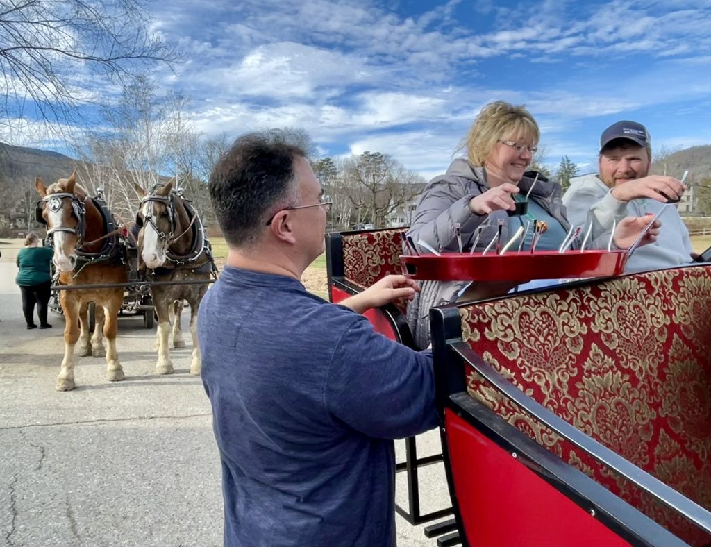 A man offering a platter of chocolate treats to a couple sitting in a horse-drawn sleigh.