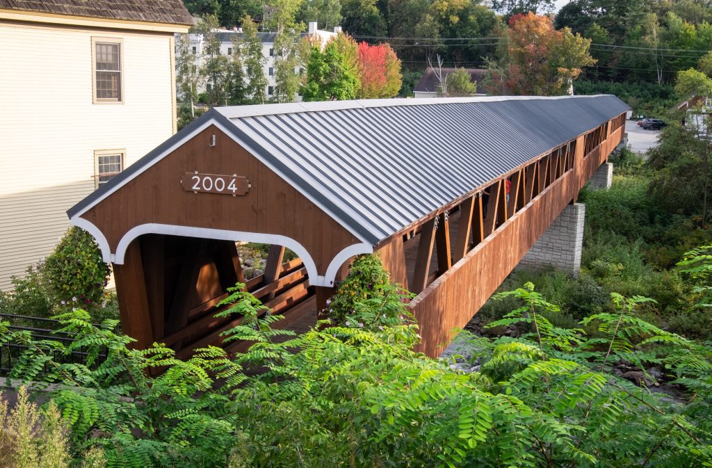 A long wooden covered bridge spanning a river, surrounded by greenery.