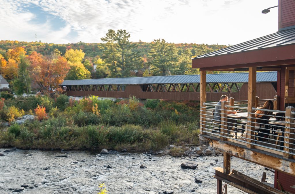 A balcony overlooking a rushing river with a long wooden covered bridge and fall colors.